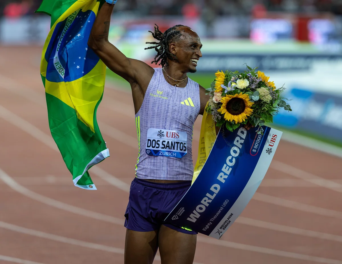 Alison dos Santos sorri e ergue a bandeira do Brasil na pista, com um buquê de girassóis e uma faixa com a inscrição World Record