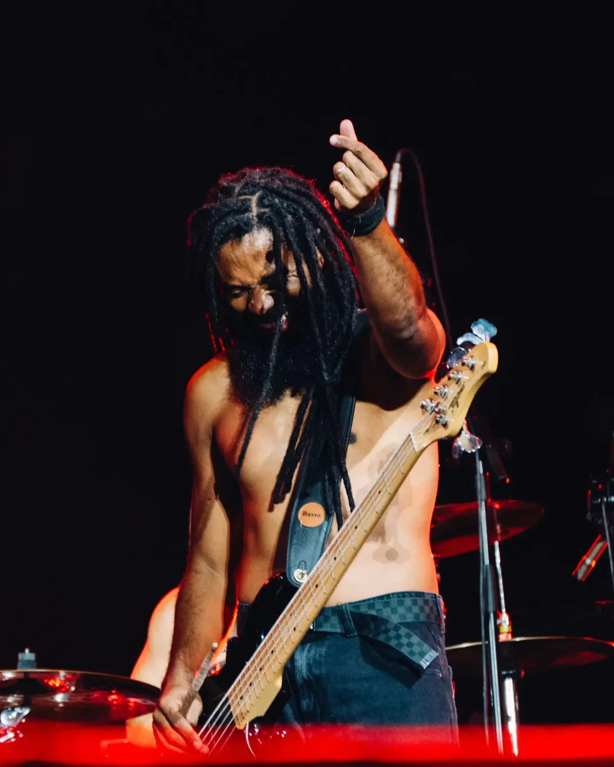 Integrante do Black Pantera toca baixo sem camisa, de dreads, com o braço erguido, sob luz vermelha