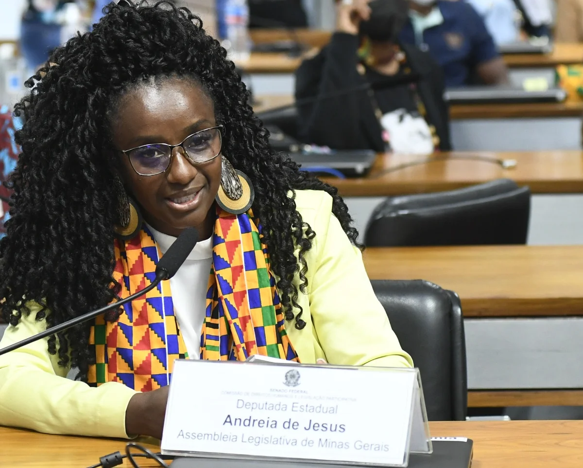 Andréia de Jesus, de óculos e lenço de estampa colorida, fala ao microfone numa mesa do Senado, atrás de uma placa com seu nome