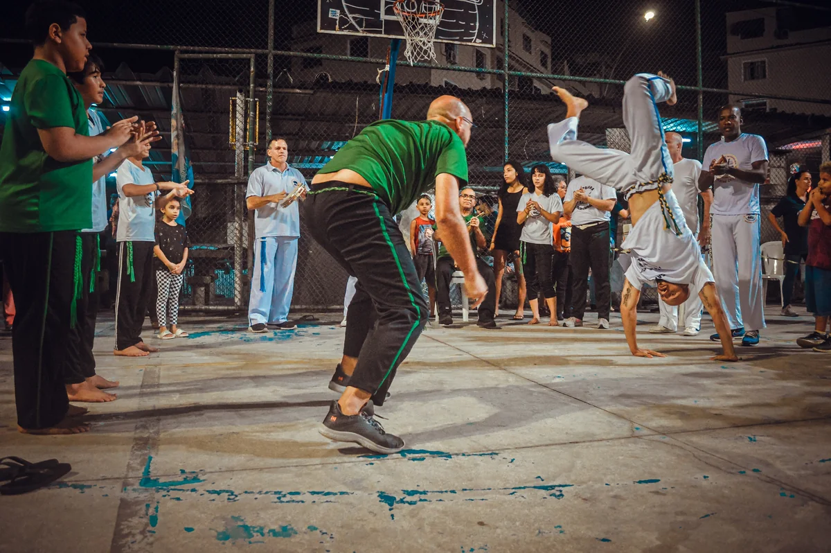 Roda de capoeira &agrave; noite, com um capoeirista de cabe&ccedil;a para baixo e outro agachado, cercados por p&uacute;blico