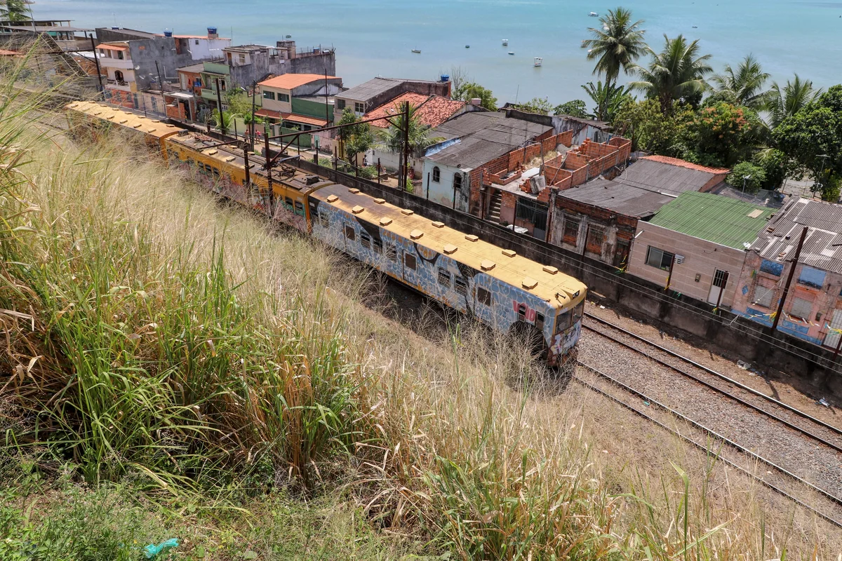 Trem do Sub&uacute;rbio passando pelo bairro de Escada, com casario colado aos trilhos e a Ba&iacute;a de Todos-os-Santos ao fundo