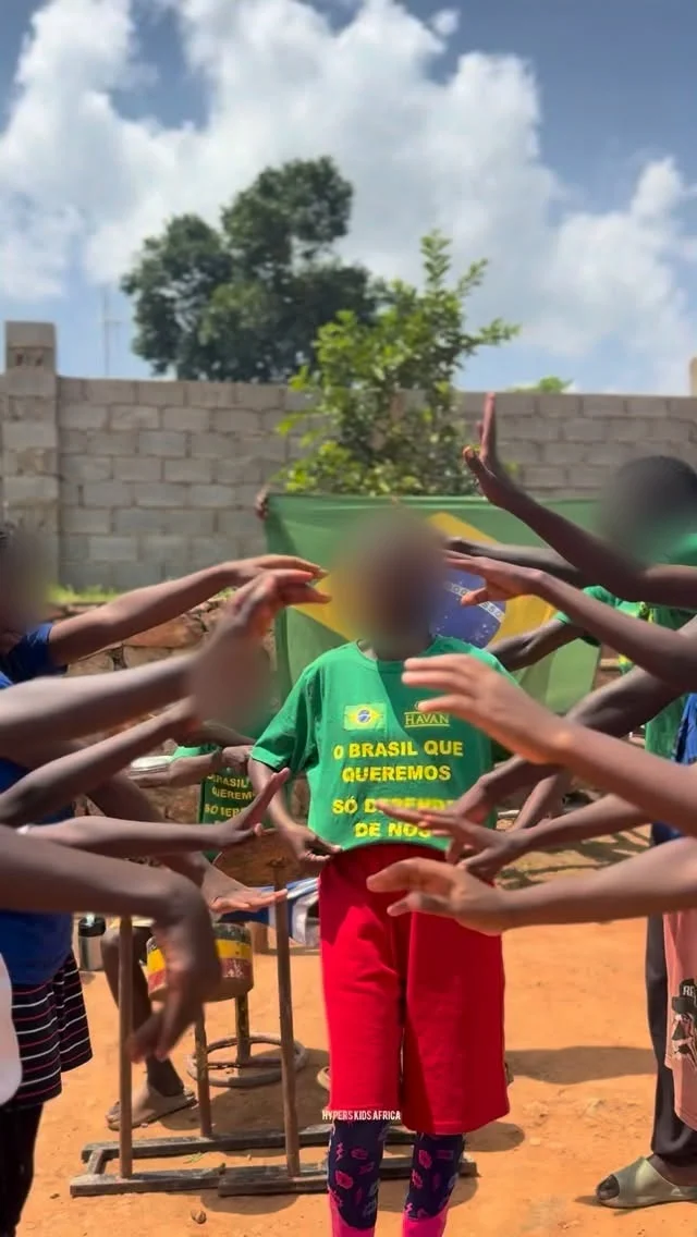 Crianças com camisetas verdes e uma bandeira do Brasil ao fundo, com os rostos borrados, fazem um corredor de mãos em Uganda