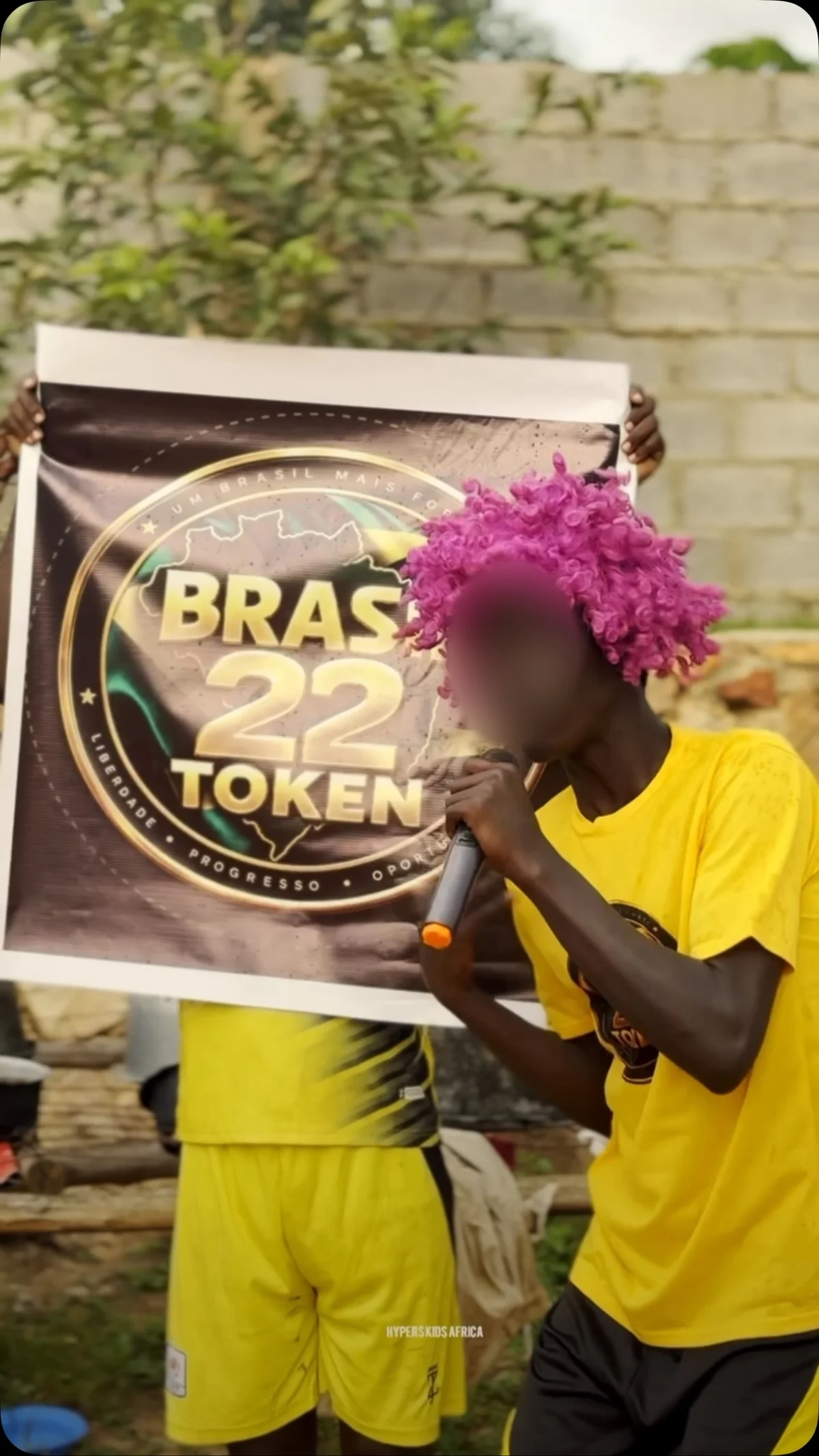 Jovem de peruca rosa, com o rosto borrado, canta ao microfone ao lado de um cartaz do Brasil 22 Token, em Uganda