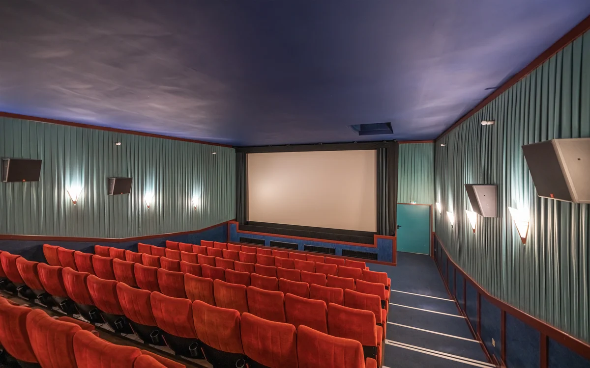 Sala de cinema vazia, com poltronas vermelhas e a tela branca acesa