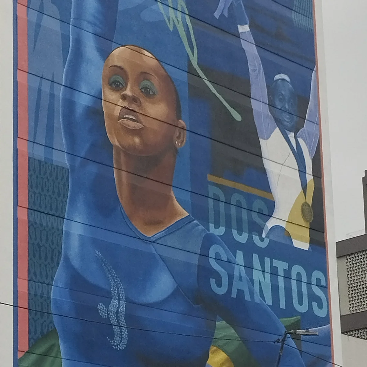 Mural pintado na lateral de um predio mostra Daiane dos Santos de collant azul com o braco erguido, ao lado das palavras DOS SANTOS