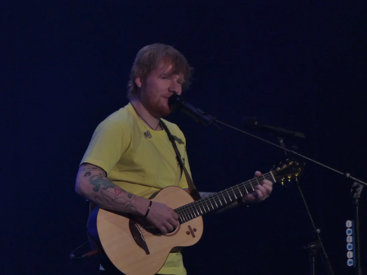 Ed Sheeran de camiseta, tocando violão e cantando ao microfone num palco de estádio