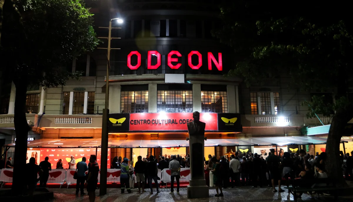 Fachada do Cine Odeon à noite, com o letreiro vermelho aceso e público na calçada da Cinelândia, no Centro do Rio