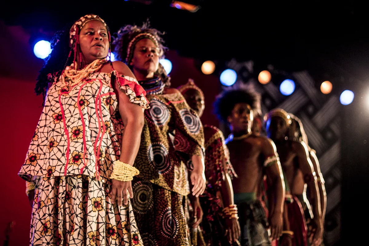 Atrizes e atores do Bando de Teatro Olodum em cena, com roupas de estampas africanas, sob luz de palco