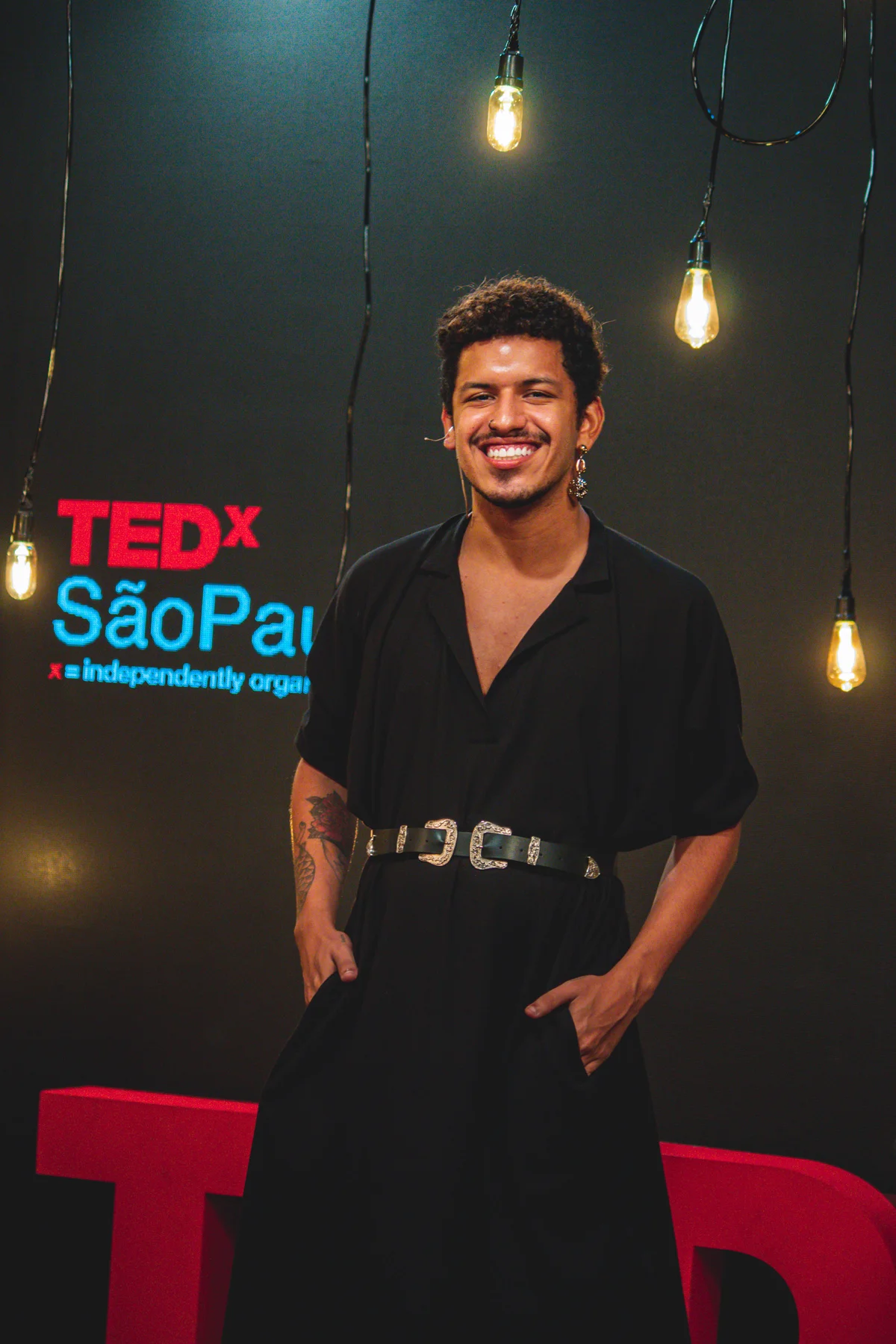 Spartakus Santiago sorri no palco do TEDxSaoPaulo, vestido de preto