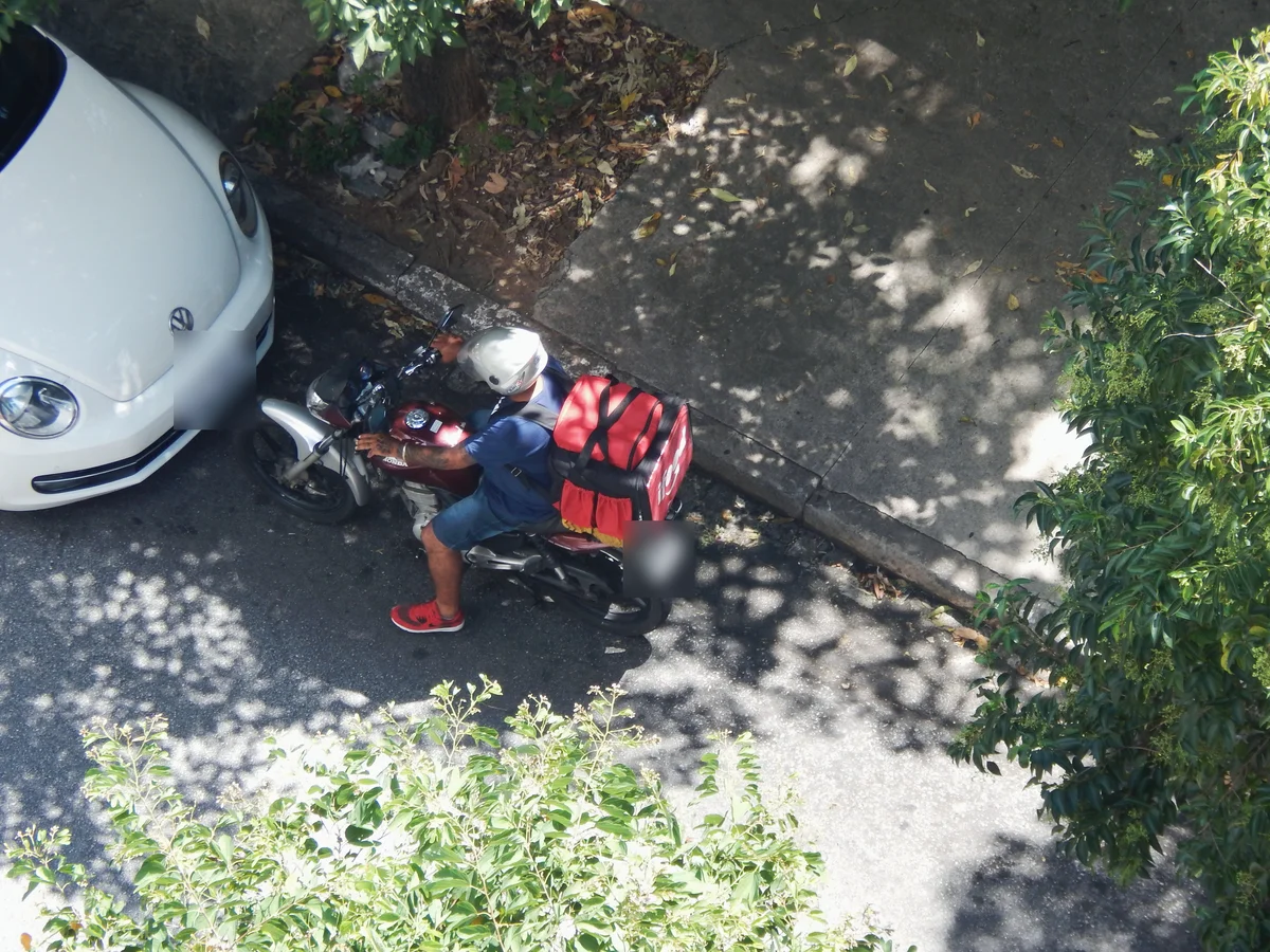 Entregador de moto com bolsa térmica vermelha, visto de cima, parado junto ao meio-fio