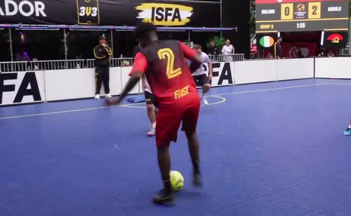 Jogador de Angola, com a camisa vermelha número 2, domina a bola numa quadra de futebol de rua em Utrecht, com o placar Irlanda 0 x 2 Angola ao fundo