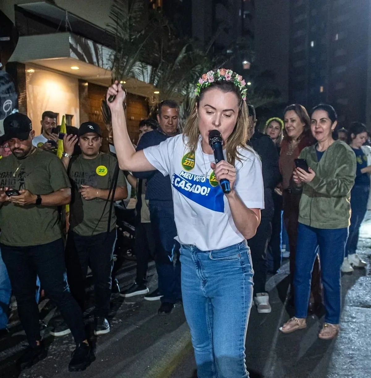Júlia Zanatta, de tiara de flores e camiseta branca, canta ao microfone com o braço erguido diante de apoiadores, à noite, numa rua de Santa Catarina