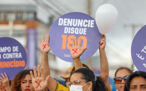 Manifestação contra o feminicídio, com cartazes do Disque 180 erguidos por mulheres com as mãos pintadas de vermelho