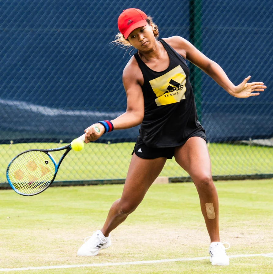 Naomi Osaka em quadra, de boné vermelho, no momento de uma devolução de forehand