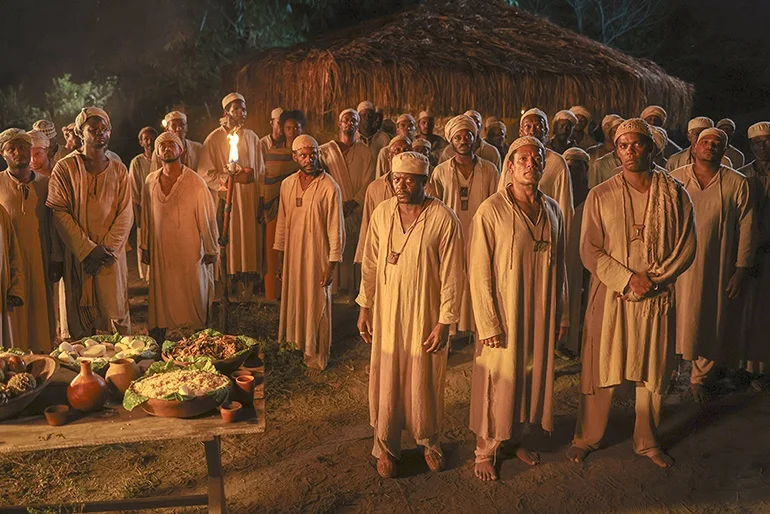 Cena noturna de Malês: dezenas de homens em túnicas claras e toucas reunidos diante de uma casa de palha, um deles segurando uma tocha
