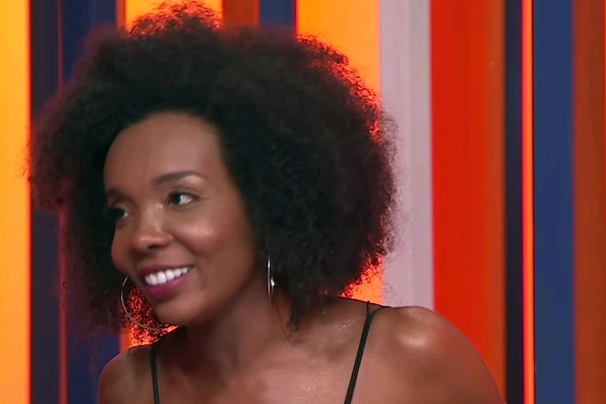 Thelminha, de cabelo black power e brinco de argola, sorrindo em estúdio de televisão