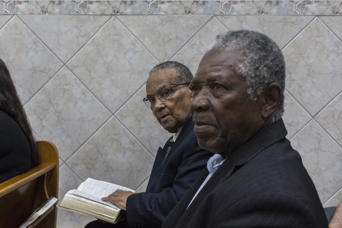 Dois homens negros sentados num banco de igreja evangélica, um deles com a Bíblia aberta