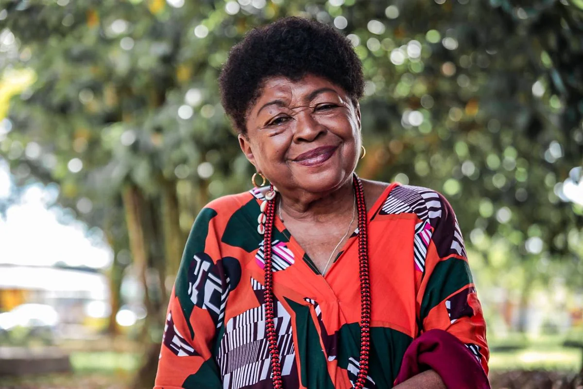 Z&eacute;lia Amador de Deus sorri, de blusa estampada e colar de contas, em uma &aacute;rea arborizada
